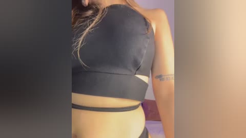 Video of a woman with light skin wearing a black, sleeveless crop top and black underwear, standing in a dimly lit room with a blurred background.