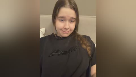 Video of a young, fair-skinned girl with long brown hair in a braid, wearing a black T-shirt, seated in a beige chair in a dimly lit room with blurred edges.