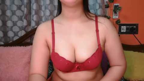 Video of a light-skinned woman with long dark hair, wearing a red lace bra that accentuates her large breasts. She is sitting on a pink sofa with patterned cushions, in a room with orange walls and a grey circular-patterned curtain.