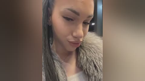 A close-up video of a woman with medium skin tone, wearing a fur-trimmed jacket, black earrings, and subtle makeup, looking down thoughtfully. The background is blurred and dark.