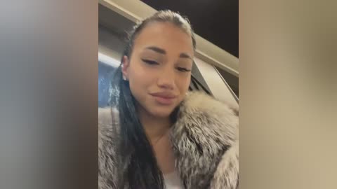 A video of a young woman with light brown skin, wearing a white shirt and a fur coat, standing in a modern interior with beige walls and a white ceiling.