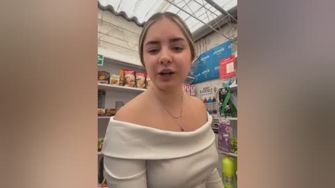 Video of a young Caucasian woman with light brown hair, wearing an off-shoulder white top, standing in a well-lit grocery store with shelves full of products.