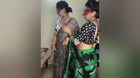 Media: Video of two women in sarees with red glasses, standing in a simple, beige-walled room. One wears a black polka-dotted saree, while the other dons a floral green saree.