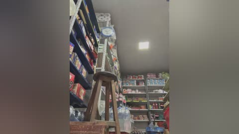 A video of a small, dimly lit convenience store with shelves filled with packaged snacks, drinks, and household items. A single wooden stool is visible in the center, and a person wearing a red shirt is partially visible in the background.