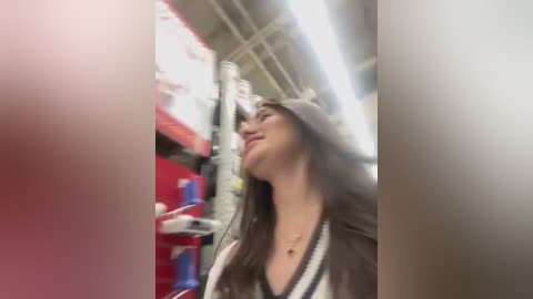 Video of a young woman with long brown hair wearing a gray hoodie, smiling while looking up at a ceiling in a brightly lit, red and white store.