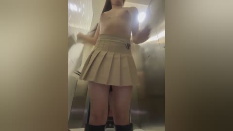 Video of a young woman with fair skin, long dark hair, and small breasts, wearing a beige pleated skirt, standing in a dimly lit, narrow corridor.
