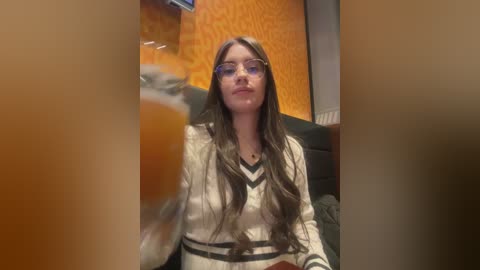 Video of a young woman with long brown hair, wearing glasses and a cream-colored sweater, sitting in a dimly lit, retro-style diner with orange patterned walls and a wooden booth.