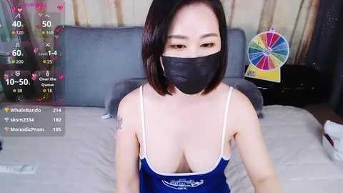Video of an Asian woman with short black hair, wearing a blue tank top, black face mask, and a small tattoo on her left arm, sitting on a bed in a dimly lit room.