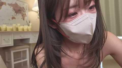 Media: Video of an Asian woman with long black hair, wearing a white surgical mask, and a delicate necklace, seated indoors, looking slightly to the right.