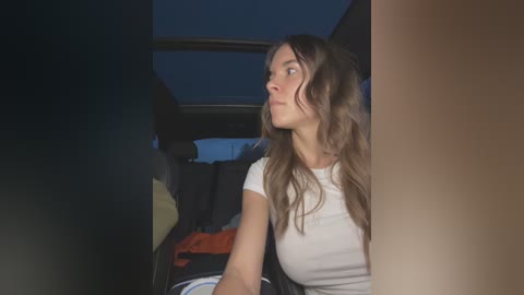 Media: Video of a young woman with wavy brown hair, wearing a white T-shirt, sitting in a car at night, looking contemplatively out the window, with a blurred view of the car interior and night sky visible through the window.