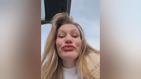 Media: A video of a young woman with long blonde hair, fair skin, and full lips, puckering them in a playful manner. She wears a white top. The background is blurred, suggesting an indoor setting.