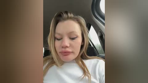 Media: Video of a Caucasian woman with light skin and blonde hair, wearing a white top, sitting in a car with gray interior, eyes closed, looking serene.