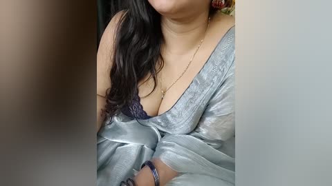 Video of a woman with long, dark hair, wearing a silver satin robe, exposing a dark lace bra, revealing cleavage. She has a gold necklace and a blue bracelet on her wrist.