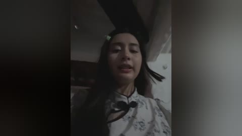 A video of a woman with long dark hair, wearing a white robe with black floral patterns, looking serene and slightly smiling, set in a dimly lit room with a dark curtain partially obscuring the scene.