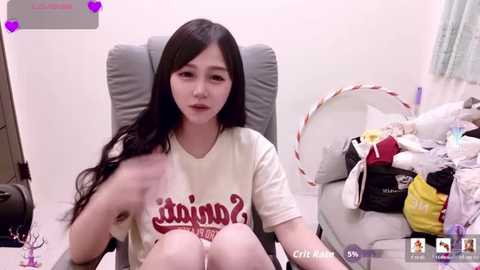 Media: Video of an East Asian woman with long black hair, wearing a cream T-shirt with Japanese text, seated on a grey chair in a cluttered room.