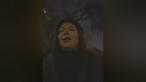 Media: A dimly lit video of a smiling woman with long, dark hair, wearing a black hat, surrounded by blurred, shadowy trees.