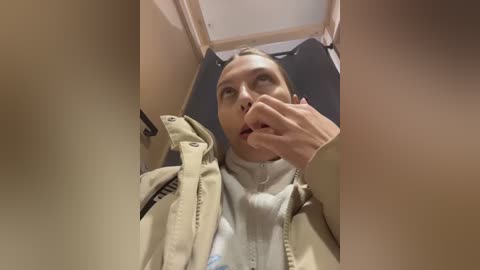 A video captures a woman in a beige jacket and white shirt lying on a hospital bed, with a concerned look on her face. Her hand covers her mouth.