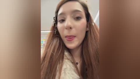 Media: Video of a young woman with long, straight, light brown hair, wearing a beige sweater. She has fair skin, light makeup, and is looking directly at the camera. The background is out of focus, showing a white wall and blurred objects.