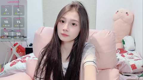 Media: Video of a young Asian woman with long, straight brown hair, wearing a striped top, lying on a bed with pink pillows and a stuffed whale. Background includes a window with blinds and a calendar.