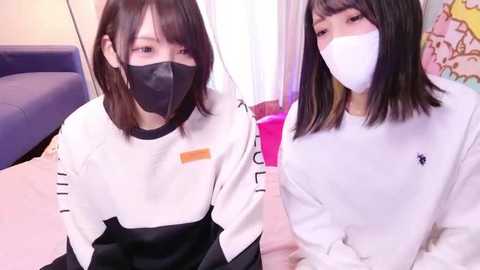 Two young Asian women, wearing white lab coats, black masks, and white face shields, pose indoors with pink and blue walls.