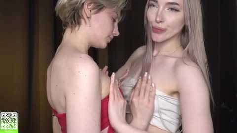 Media: Video of two young women with fair skin, one with short blonde hair in a red dress, the other with long blonde hair in a white bandeau, embracing affectionately.