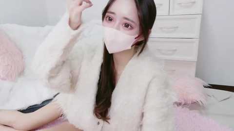 Video of an East Asian woman with long black hair, wearing a pink face mask, white fuzzy robe, and light pink socks, sitting on a white bed with fluffy pillows. Background shows a white dresser.