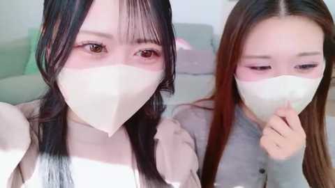 Video of two East Asian women with long black hair, wearing white face masks, one holding her finger to her lips, in a light-colored room with blurred background.