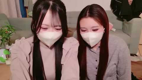 Media: Video of two East Asian women with long black hair, wearing white face masks, beige sweaters, and sitting close together on a light gray couch. Background features a potted plant and a person partially visible on the right.