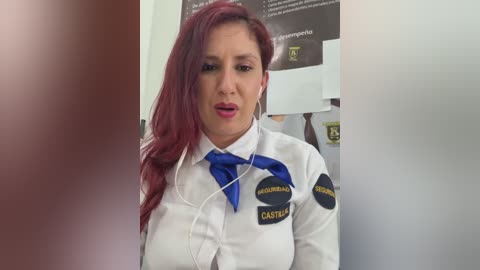 A video of a middle-aged woman with long, straight red hair, wearing a white shirt with blue accents and a blue tie. She appears to be in a professional setting, with documents and a computer screen in the background.