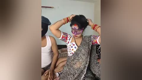 Video: A woman with dark hair and a colorful mask, adorned with red and gold bangles, sits on a bed, being assisted by another person in a simple room.
