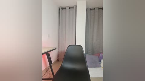 Media: Video of a minimalist bedroom with a black chair in the foreground, a white bed against gray curtains, and a small white table to the left.