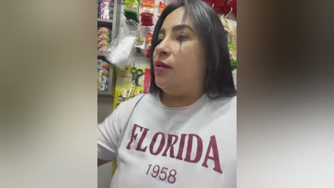 Video of a Latina woman with medium skin tone and shoulder-length black hair, wearing a white Florida State University shirt, standing in a colorful convenience store with shelves of snacks and drinks in the background.