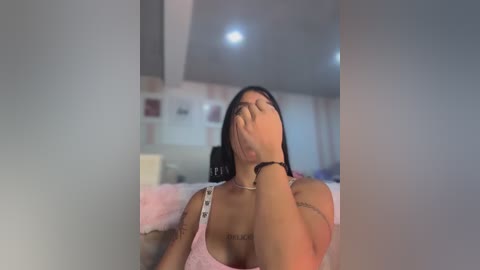 Media: Video of a Latina woman with medium skin tone, wearing a pink bralette, lying on a bed, covering her eyes with her hand, and holding a phone.