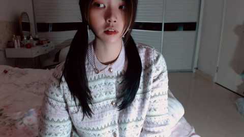 Media: A video of a young Asian woman with long black pigtails, wearing a patterned sweater, sitting on a bed in a dimly lit bedroom with a vanity table and closed window blinds.
