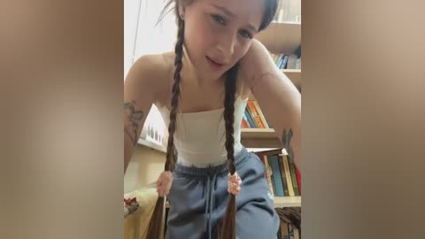 Video of a young woman with long braided hair, wearing a white tank top and blue pajama pants, leaning on a wooden chair in a cozy, book-filled room.