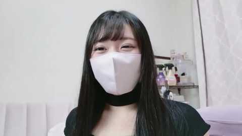 Video of an Asian woman with long black hair, wearing a white face mask, black choker, and black top, standing in a modern bathroom with a white curtain and shelves filled with toiletries.