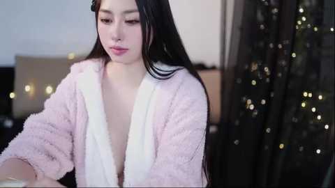Video of a young Asian woman with long black hair, fair skin, and a slender physique, wearing a fluffy pink robe. Background includes a dark curtain and festive lights.