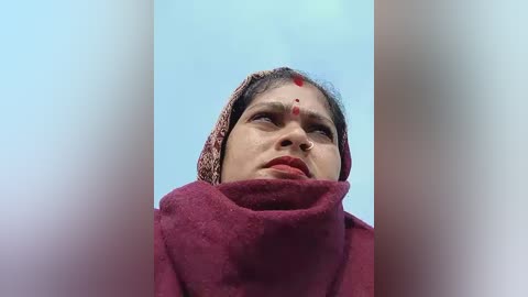 Media: Video of a woman with a fair complexion, wearing a maroon shawl, and a traditional headscarf with a red bindi on her forehead, set against a soft, gradient blue sky background.