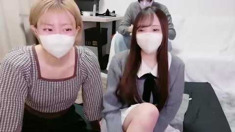 Video of two young Asian women with long hair and white masks, wearing checkered shirts and skirts, sitting on a black floor in a white room, with a person in the background.