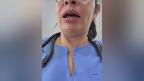 Media: Video of a light-skinned woman with glasses, wearing a light blue scrub top with a V-neckline and a stethoscope around her neck, speaking with her mouth open, in a blurred indoor setting.