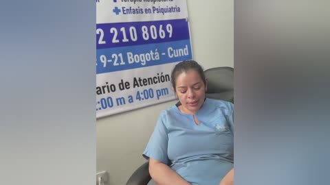 Media: Video of a middle-aged woman with light skin, brown hair tied back, wearing light blue scrubs, sitting in a hospital chair, in a room with a blue and white emergency phone number sign.
