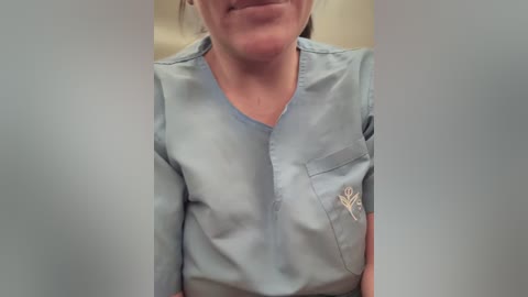 Media: Video of a woman in a light blue scrub top, featuring a small emblem on the left pocket. Her face is partially visible, smiling with closed lips, and her brown hair is tied back. Background is blurred, likely an indoor setting.