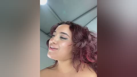 Video of a young woman with medium brown skin and long, wavy, dark auburn hair, wearing black eyeliner and light makeup, smiling, taken indoors with a white ceiling and visible light fixture.