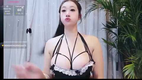 Video of an Asian woman with long black hair, wearing a revealing black and white lingerie with a crisscross top, standing against a backdrop of sheer curtains and greenery.