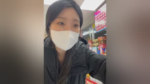 Video of an Asian woman with long black hair, wearing a white mask and black jacket, looking into a grocery aisle filled with brightly colored packaged goods.