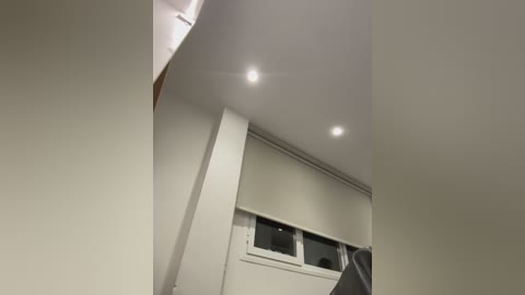 Video of a modern, minimalist room with smooth, white walls and ceiling. Three recessed lights illuminate the space, highlighting a window with closed beige blinds. The room's clean lines and simple decor create a serene, uncluttered atmosphere.