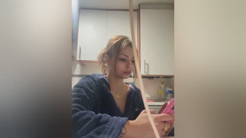 Video of a young woman with light skin, wearing a blue robe, sitting in a modern kitchen, focused on a pink phone.
