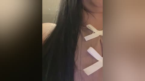 Video of a person with long, straight black hair, wearing a beige sweater with white X-shaped tape over the chest, partially blurred background.