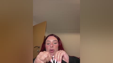 Media: A video shows a woman with red hair and fair skin, wearing glasses, sitting with her eyes closed and hands clasped, in a beige room with a wooden door.