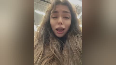 Video of a young woman with long, wavy, light brown hair, slightly parted lips, and closed eyes, wearing a casual outfit, blurred background.